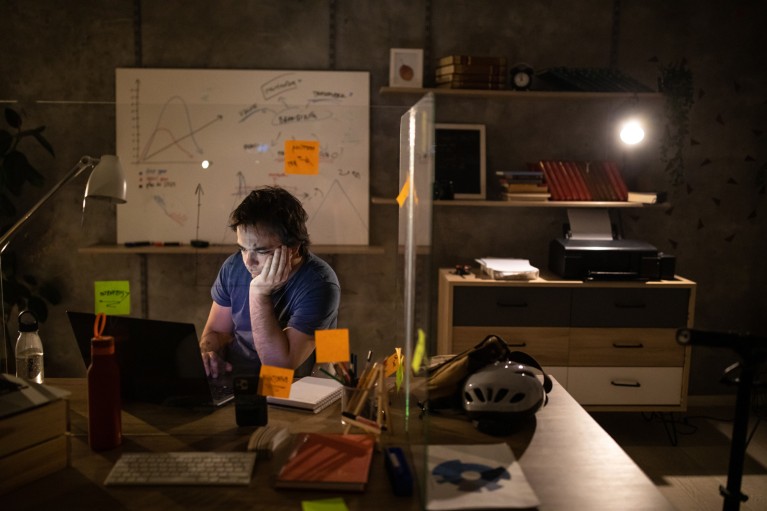 A young man sitting at a desk working on a laptop in a dark office with a whiteboard behind him with charts written on it