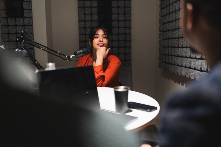 Млада жена седи за столом са микрофоном, замишљено слуша колегу током сесије снимања подкаста у професионалном студију