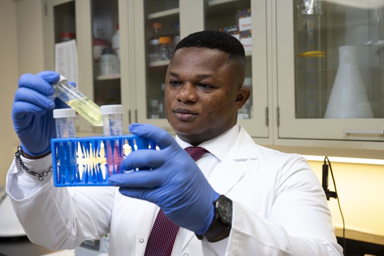 Thomas Karikari wearing white coat and blue gloves, holding laboratory equipment in both hands. He looks towards a plastic tube containing liquid.