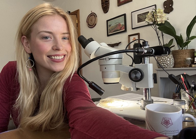 A blonde woman smiles while sitting next to a microscope