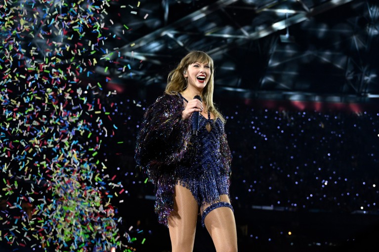 Taylor Swift smiles while standing on stage during a concert in a large arena with multi-coloured confetti falling behind her