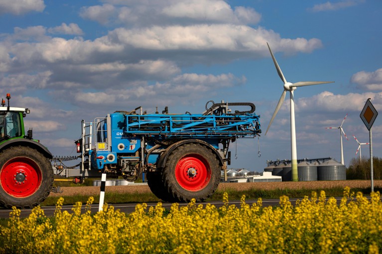A field of yellow flowering rapeseed. In the middle distance, a tractor pulls a sprayer past wind turbines.