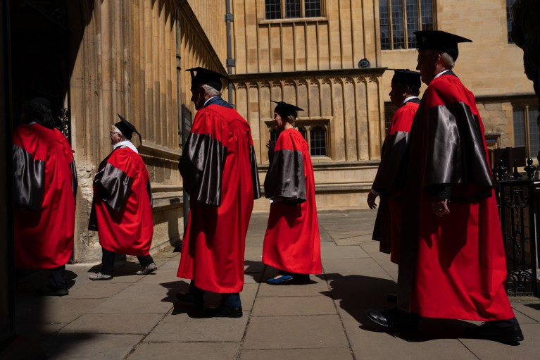 People wearing red and black academic gowns and mortar boards process across a sunny quadrangle into a shadowy doorway