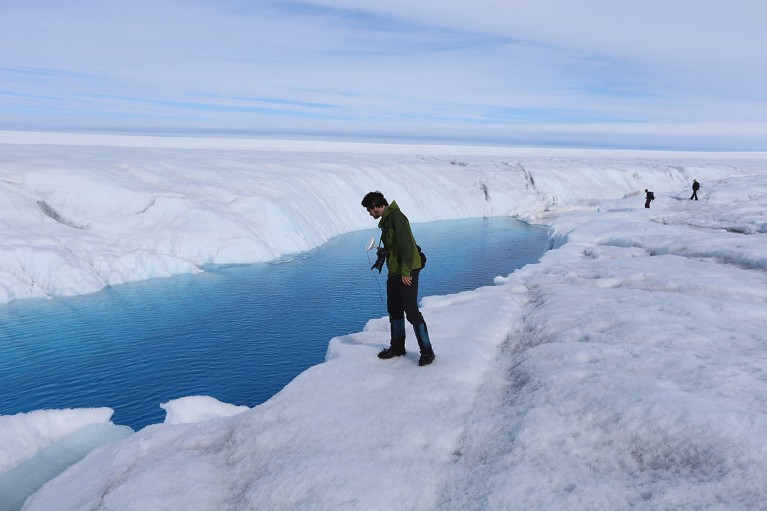 A Ph.D. student standing on the glacial ice sheet in Greenland looks down at a blue meltwater stream.