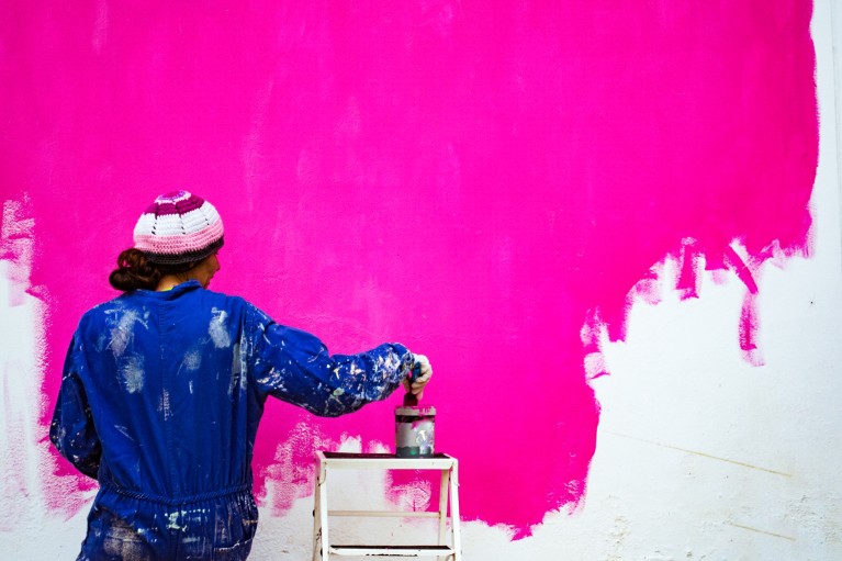 Standing beside a wall half painted in bright pink, a woman in blue work overalls covered in paint putting a paint brush into a paint tin balanced on a step ladder