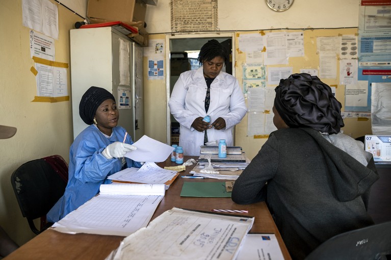 A woman talks to one health-care worker wearing a protective gown and another wearing a lab coat.