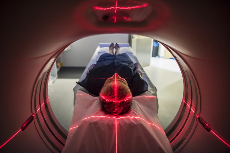 At centre, a person lying on a bed entering a scanner. A cross of red light shines on the top of their head.