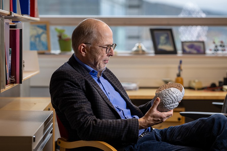 Jan Haavik sits in a chair, looking at a model of the human brain. Behind him are shelves with books on them.