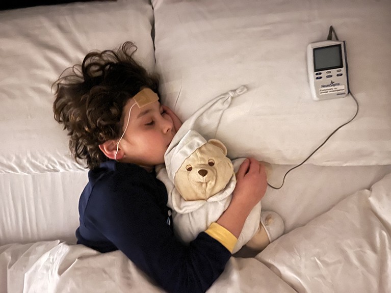 View from above of child with eyes closed lying in bed, one arm around a teddy bear. A sensor is attached to the child's forehead, connected by a wired to a device on a pillow on the bed.