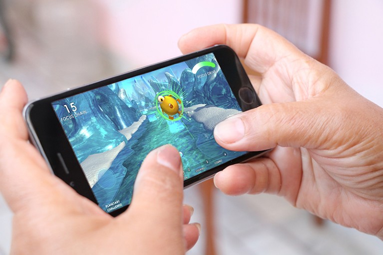 Close-up of two hands holding a phone. The two thumbs are touching the screen. The background of the screen is blue, green, and white. To the upper right of the phone screen is a round, yellow object with what looks like an eye, mouth, and arms and legs.