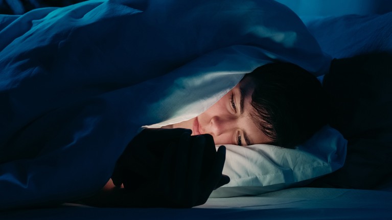 To the right, face of boy lit up by the phone in his hands. He is lying in bed covered by duvet, head on pillow.