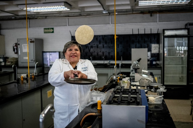 Wearing a white lab coat while standing in a laboratory, Raquel Gomez Pliego holds a frying pan in two hands and flips a tortilla into the air