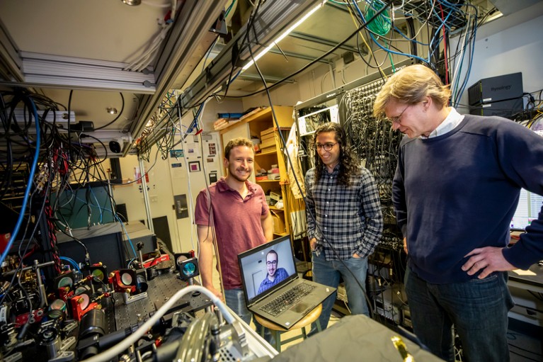 Three researchers standing around a laptop with another person on the screen, in a lab filled with experimental equipment and tangles of wires.