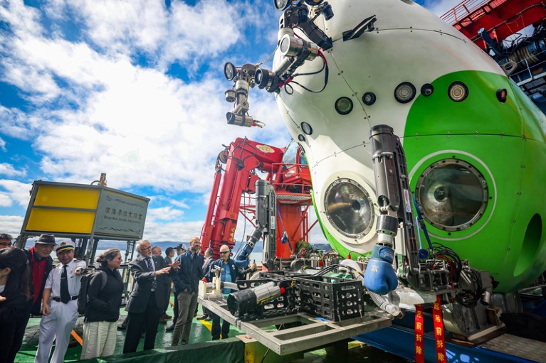 On the deck of a ship, a crowd of people look up at a suspended white and green submarine, featuring large lights and robotic arms.