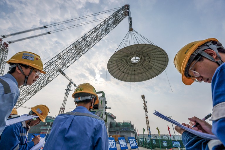 Workers in blue overalls and yellow hard hats look on and make notes as a huge dome is hoisted into the air by a crane at a construction site.