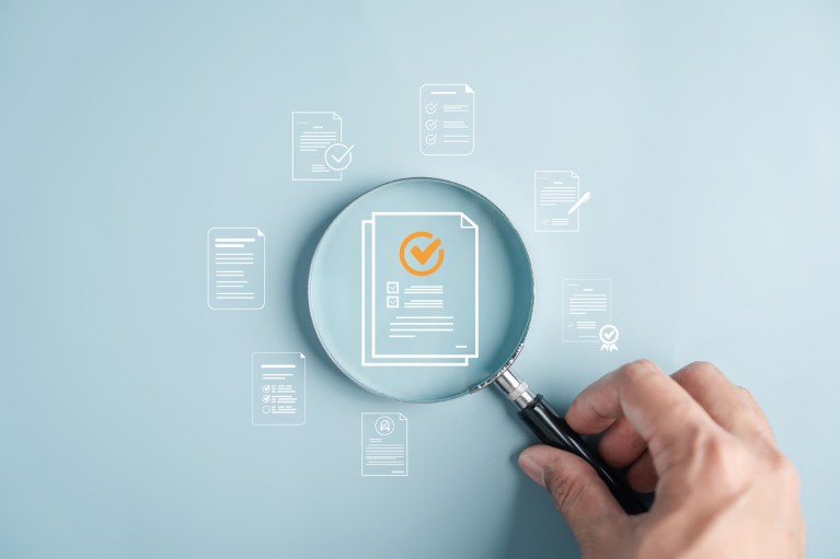 A hand holding a magnifying glass over a light blue flat surface covered with icons representing computer documents, the magnifying glass hovers over a document with an orange tick