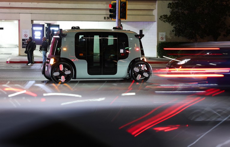 An electric autonomous robotaxi waits to make a left turn at a traffic light, while other cars zoom past.
