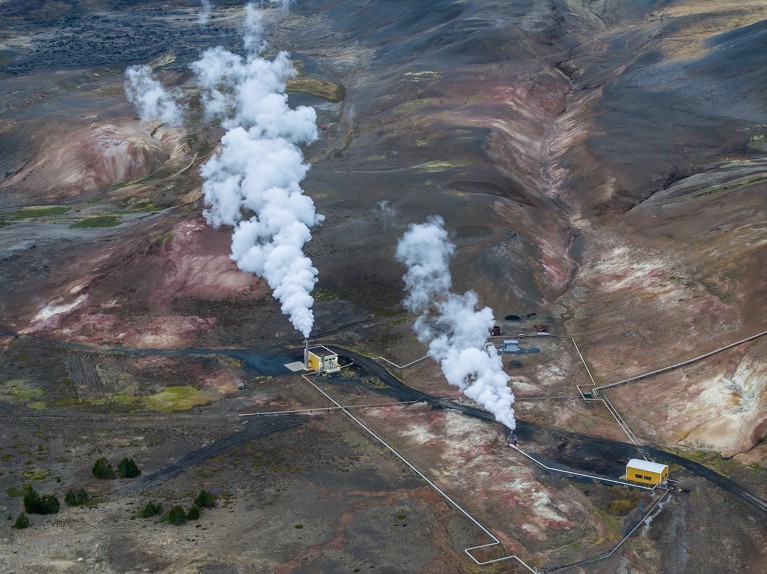 Aerial view of two jets of steam emerging from industrial buildings among low red hills.