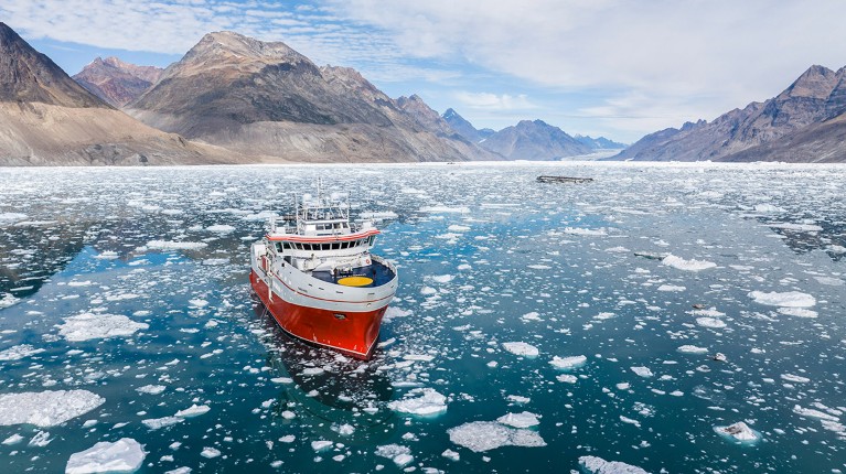 An aerial view of the Tajaroq research vessel navigating Greenland's icy costal waters with mountains in the background.