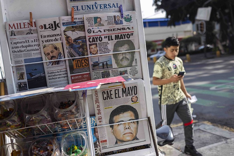 A newspaper stand displays multiple newspaper front pages. Next to the stand, a person walks by holding a mobile phone.