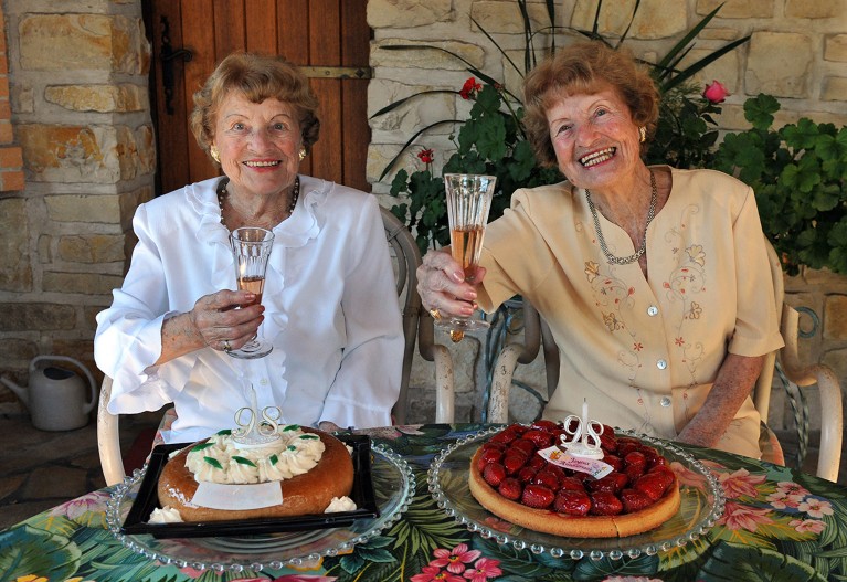 Smiling twin people are seated at an outdoor table set with two cakes and face the camera while raising their champagne flutes in a toast.