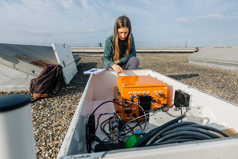 Person working on a rooftop. In front is a white open container with an orange electronic device and various cables, and a backpack placed nearby.