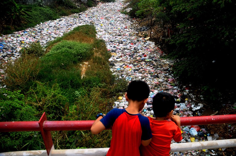 Двоје деце гледано отпозади гледају преко ограде моста у гомилу пластичног смећа која зачепљује реку, са дрвећем на обали у Западној Јави.