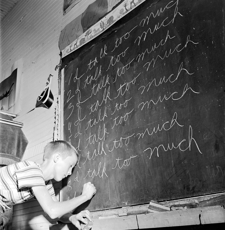An elementary school student writing the same sentence repeatedly in chalk on a chalkboard from the mid-20th century.