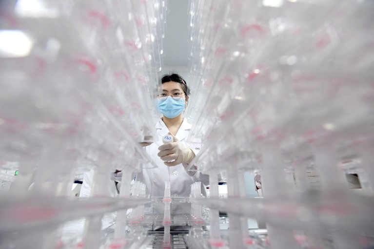 A laboratory technician in PPE, seen between two stacks of samples in tubes.