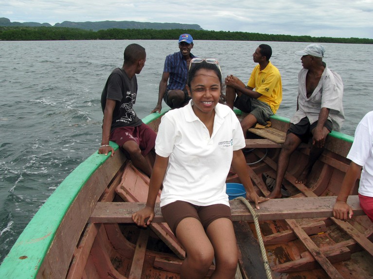 Ando Rabearisoa sits near the center of a small boat with five other people and a shoreline of trees in the background