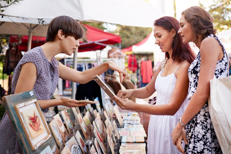 At an open-air market stall, a vendor in discussion with two customers points out a detail of a small painting for sale.