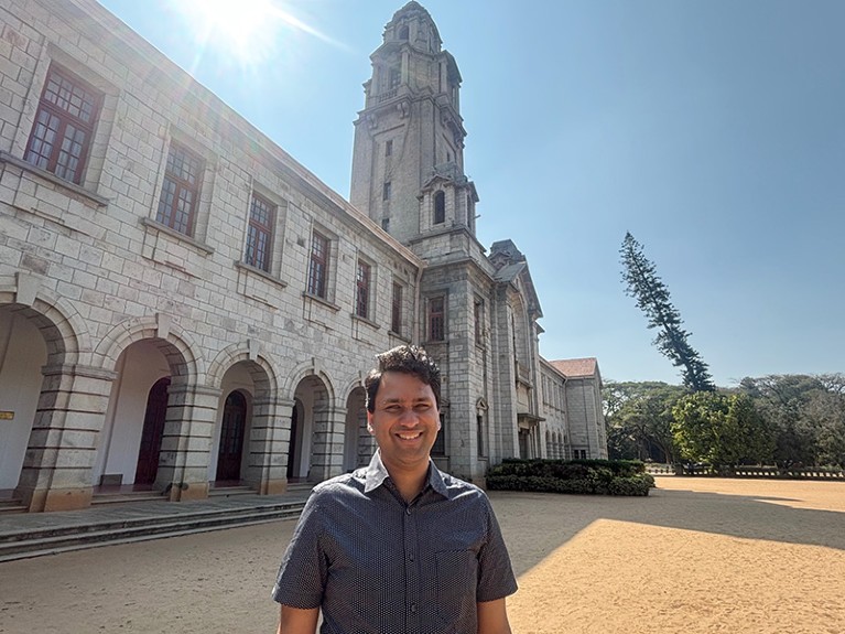 Ananth Govind Rajan stands outside of the Indian Institute of Science in Bengaluru, India. The sun shines brightly in the sky behind the ‘cathedral like’ silver-grey granite tower and buildings.