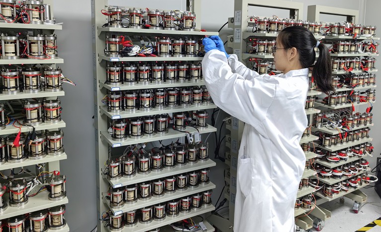 Xiaona Li stands in front of a wall of hundreds of all-solid-state batteries mounted onto shelves. She wears a white lab coat and protective blue gloves as she adjusts a battery on the top shelf. The batteries are about the size of a soda can and wrapped in silver and red wire.