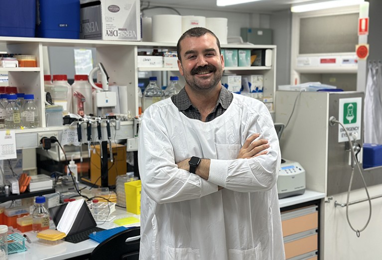 Rhys Parry stands in his laboratory in a white lab coat. He has his arms folded and is smiling at the camera.