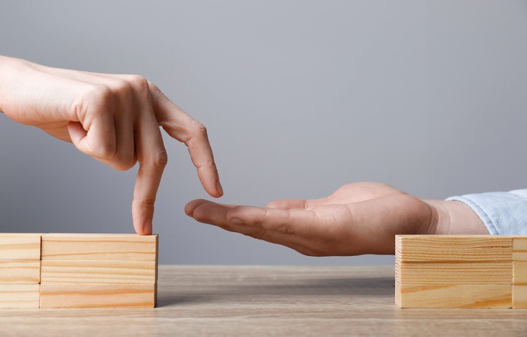 Поглед изблиза на руке двоје људи и сет дрвених блокова који илуструју особу која помаже свом партнеру да пређе недовршени мост.
