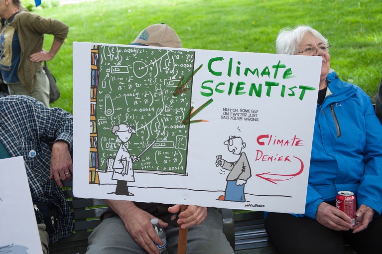 People sit on a bench, with one person holding a sign of a cartoon in support of climate change.