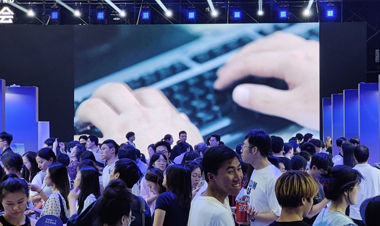 Група људи који траже посао испред великог екрана који показују руке на тастатури присуствују сајму послова како би сазнали о потражњи за вештачком интелигенцијом у технологији.