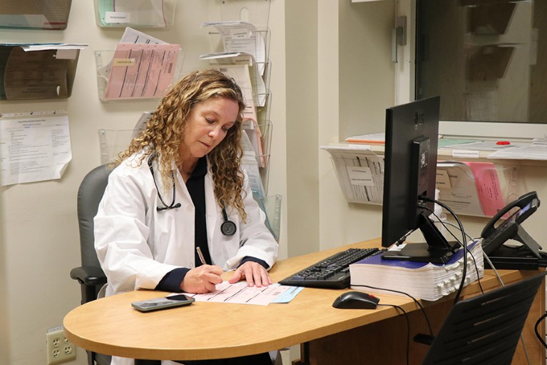 White woman wearing white coat seated at desk writing on pink and blue papers