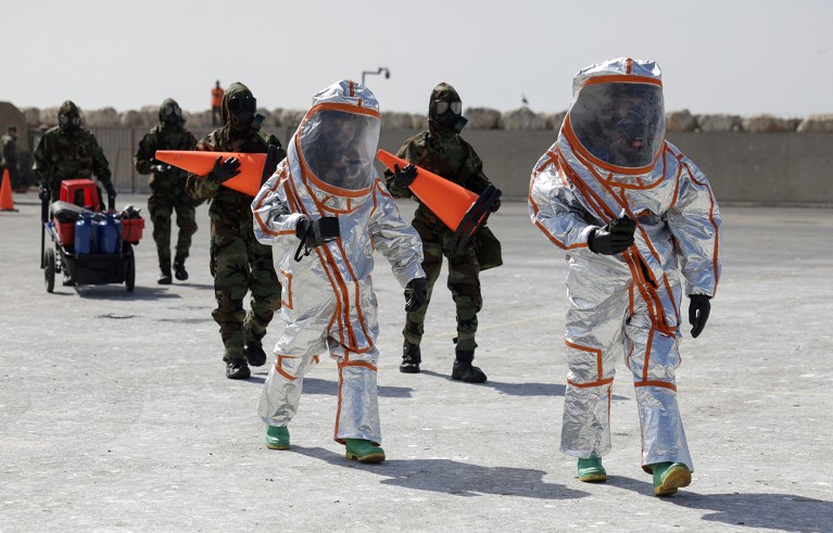 Two people in silver suit safety suits walk on a grey surface, followed by people in black protective gear holding various pieces of equipment.