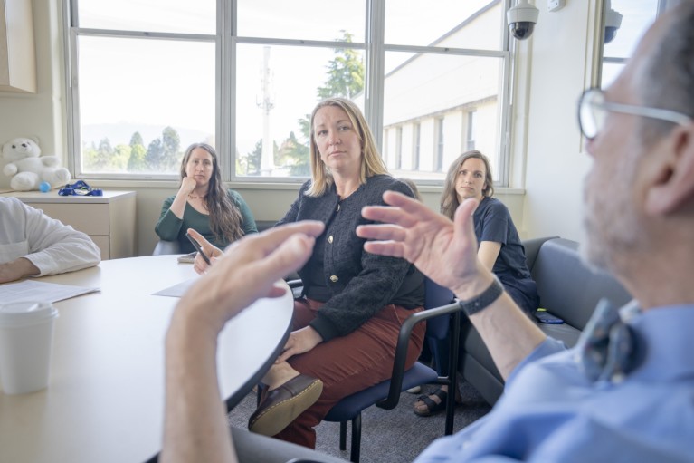 Три жене на крају овалног стола, слушају како мушкарац у првом плану говори.