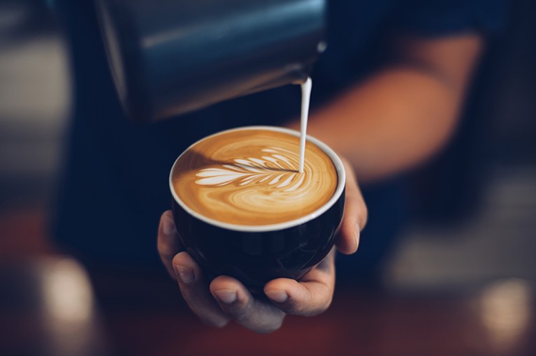 Close-up view of a person holding a cup of coffee while pouring steamed milk to create a leaf-shaped latte art pattern on the surface