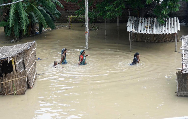 Aerial view of four people walking through waist-high water past flooded buildings..