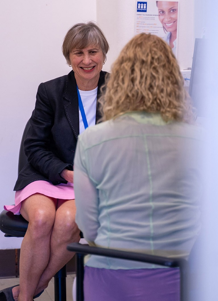 Seated white woman in a small room talking to a female patient with long curly.