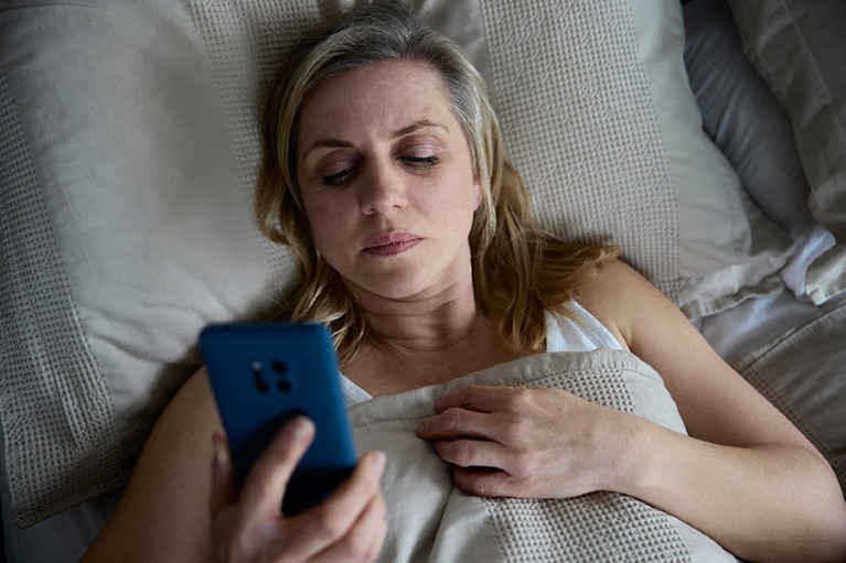 Aerial view of white, middle-aged woman in bed looking at her phone with a serious expression.