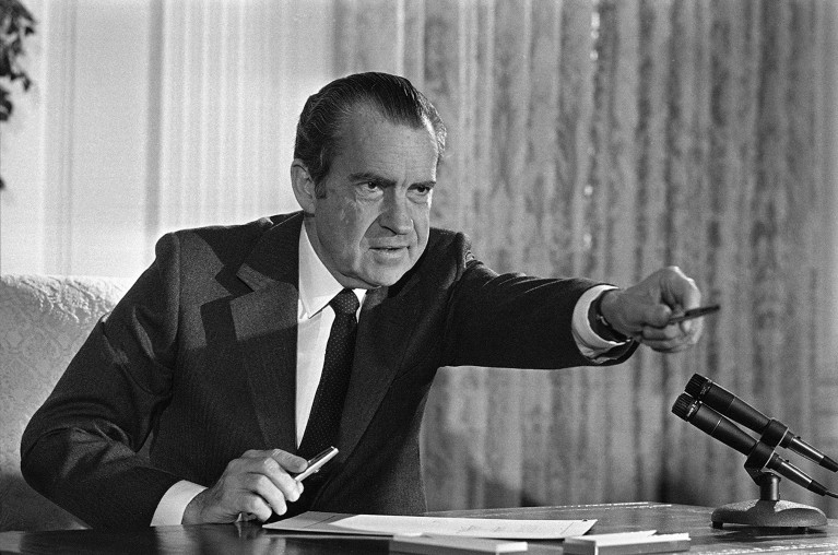Black and white photo of Richard Nixon sitting at a desk, holding a pen in one hand and offering another to someone out of frame with the other.