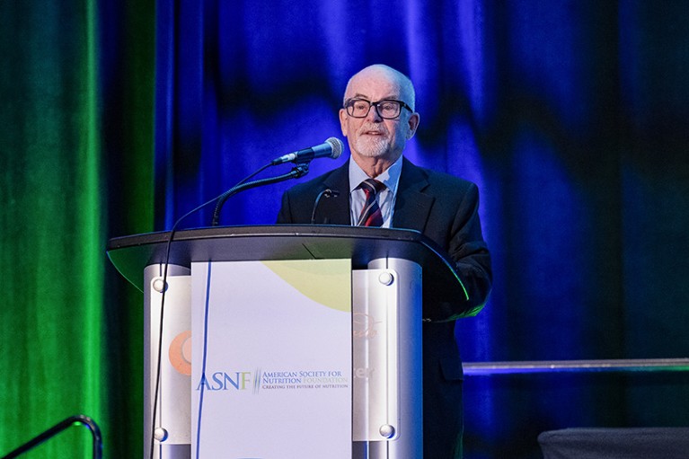 Paul Coates stands behind a podium on a stage, speaking into a microphone. The podium features a sign with the logo and text for the American Society for Nutrition. The background consists of tall curtains lit with blue and green stage lighting. Paul M. Coates is wearing a dark suit with a collared shirt and striped tie.
