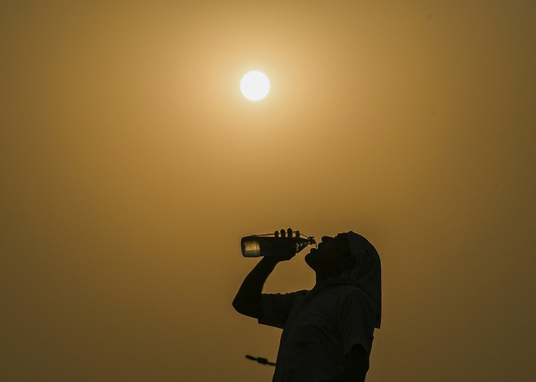 A man drinks water from a bottle on a hazy, hot day as the temperature rises, with the sun fully visible in the background.
