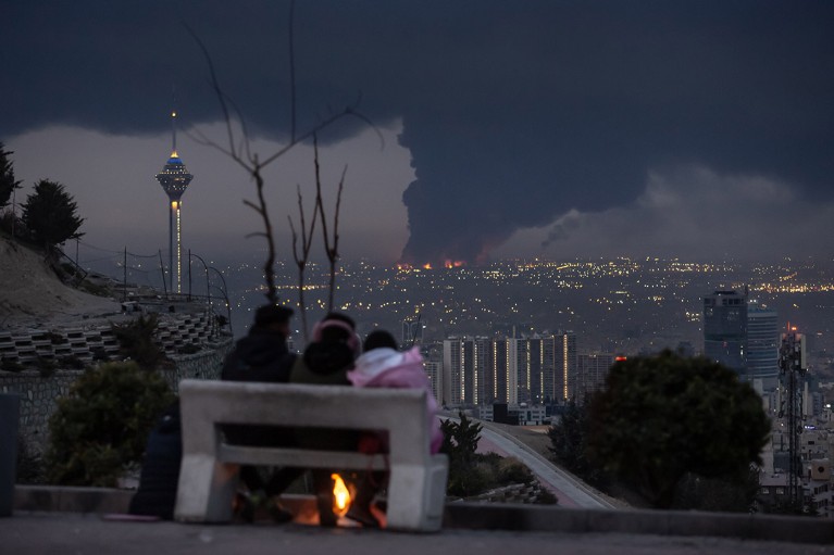 Three people sat on a bench overlook a dark Tehran skyline engulfed in black smoke rising from a burning oil depot.