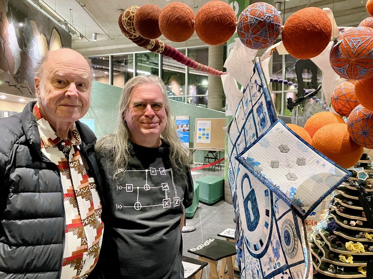 Charles H. Bennett and Gilles Brassard pose for a photograph besides a Cryptography Quilt at Mathemalchemy 2024.