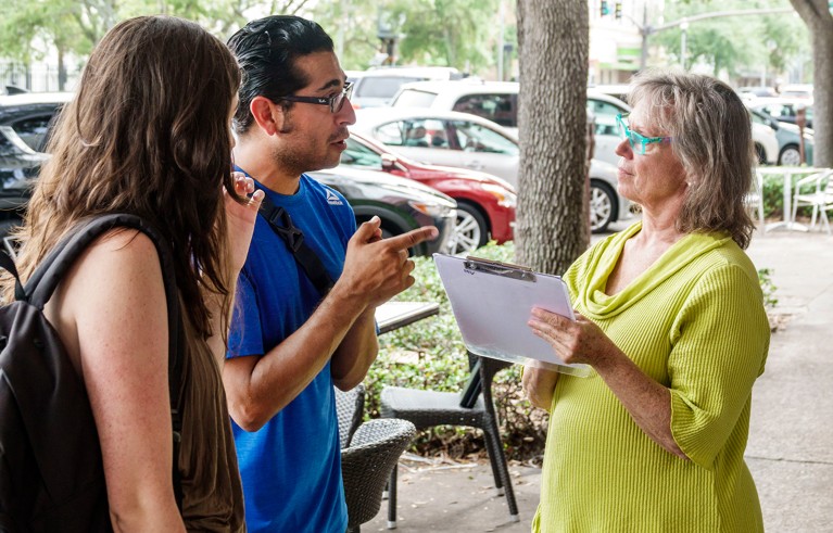 Woman with a clipboard asking two pedestrians survey questions.